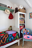 Bed with colorful bedspread, shelf, and blue rattan armchair with fabric figures in guest room