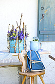 Blue pot with veronica, Delphinium, and birch twigs, in front of it a stool with a blue rucksack