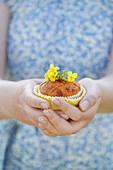 Person holding a muffin with small yellow flowers as decoration