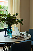 A vase of leafy branches, a laptop and a magazine on a round table