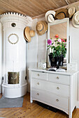 White tiled stove, mirrored chest of drawers and collection of hats in rural ambience