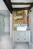 Chest of drawers with sink in light grey bathroom with partly exposed brickwork and rustic wooden beams