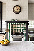 View over kitchen island with marble worktop to gas cooker in wall niche with green-white tiles, above kitchen clock