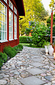 Gartenweg aus Pflastersteinen und Betonplatten am rot gestrichenem Holzhaus entlang