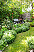 Weiß blühende Pflanzen am Rasen entlang, im Hintergrund Sitzplatz im Garten