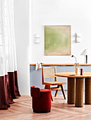 Round table with chair and red pouf seat in room with floor-length curtains