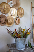 Hutsammlung an der Wand und Zinkeimer mit Blumen