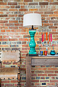 Wooden console with turquoise blue table lamp and candlestick next to leather chair in front of brick wall
