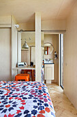 Double bed with colorful bedspread, small vanity table, mirrored sliding door behind it and view into the bathroom
