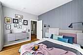 Blue and gray bedroom with pastel pink accents. A double bed with a headboard composed of several upholstered elements in different colors and fabrics. Wallpaper with vertical stripes in shades of muted blue and gray. The bed is mounted on a raised f