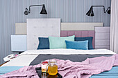 Blue and gray bedroom with pastel pink accents. A double bed with a headboard composed of several upholstered elements in different colors and fabrics. Wallpaper with vertical stripes in shades of muted blue and gray. The bed is mounted on a raised f