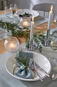 Dining table set with white plates, light blue napkins, fir branches and candle lights