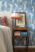 Bedside table with books and lamp in front of floral wallpaper