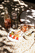 Picnic scene with bread, olives, tomatoes and drinks on a patterned blanket in the garden