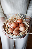 Hands holding a bowl of brown eggs on a straw base