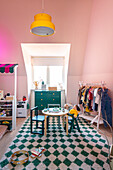 Children's room with sloping ceiling, pink walls, yellow hanging lamp and green and white carpet in harlequin pattern