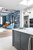 Blick über Kücheninsel auf Kühlschrank und Einbaugeräte, im Hintergrund Wohnzimmer mit blauer Regalwand