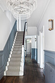 Hallway with staircase, striped runner, chandelier and wall panelling in white and grey-blue