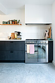 Modern kitchen unit with dark-coloured cabinet front, stainless steel appliances and wooden shelving