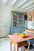 Wooden table with fruit bowl in a bright kitchen with grey display cabinet