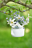 Blumenarrangement mit Narzissen und Vergissmeinnicht hängt im Stoffbeutel an einem Ast