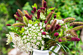 Summer flowers and grasses in white and red in a vase in front of green foliage