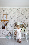 Kind spielt mit Puppenhaus an weißem Tisch in Zimmer mit floraler Tapete