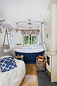 Freestanding bathtub in a small, cosy bathroom with wood-burning stove and sheepskin armchair