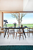 Dining area with wooden chairs and telescope in front of panoramic window with countryside view