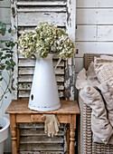 Small wooden table with enamel jug and dried hydrangeas in front of vintage shutters