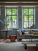 Living room with large windows, corner sofa and round side table with bouquet of flowers