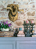 Robust wooden tray with flowers, wooden candlesticks on chest of drawers, ram's head above on natural stone wall
