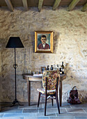 Small console table with floral chair, floor lamp and gold-framed portrait painting on natural stone wall
