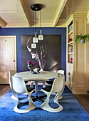 Round dining table with white chairs and large piece of art on a blue wall