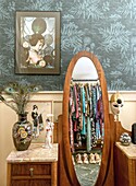 Mirror reflecting clothes rack in a room with wallpaper with botanical pattern and decorative vase