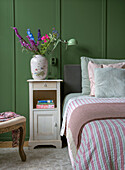 Bouquet of flowers in antique vase on white bedside table in bedroom with green wall