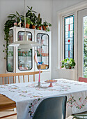 Dining table with embroidered tablecloth and plants on display cabinet