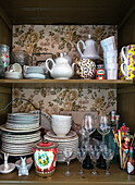 Open kitchen cupboard with various crockery and patterned wallpaper background