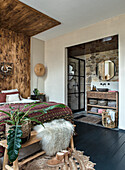 Bedroom with wooden panelled wall, black floorboards and adjoining bathroom with stone wall and round mirror