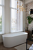 Freestanding bathtub in front of large windows with blinds and pendant lights