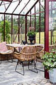 Seating area in the red glass house with rattan armchairs and wooden table on a paved floor