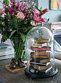 Stack of books under a glass bell on a wooden table next to a bouquet of roses and lilies