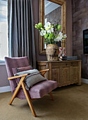 Designer armchair in shades of pink in front of antique wooden chest of drawers with ceramic vase and flower arrangement