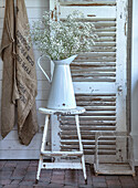 Large white jug with baby's-breath on a rustic stool in front of shutters