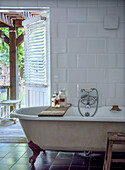 Freestanding vintage bathtub in front of white tiled wall in rustic bathroom with garden view