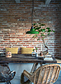 Dining area with rattan furniture, rustic wooden table and green hanging lamp in front of brick wall