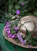 Strohhut mit rosa Wicken (Lathyrus) und violetter Spornblume (Liatris) auf grüner Bank