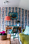 Desk with vintage lamp in front of patterned wallpaper, green sofa and wicker table next to it