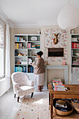 Person in front of bookshelf in living room with floral wallpapered wall