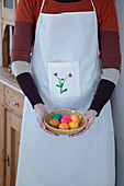 Person in apron with embroidered flower holding bowl with coloured Easter eggs